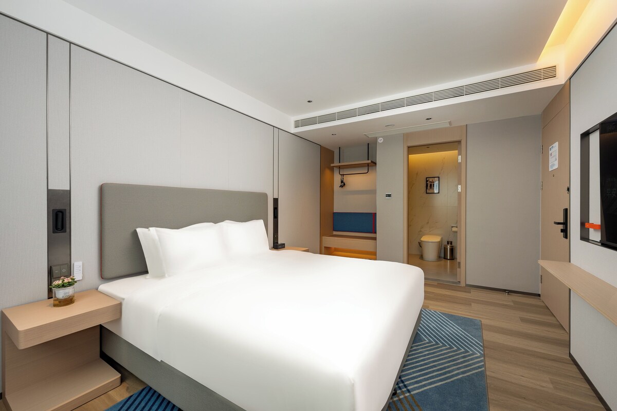 A modern hotel room with a large bed, bedside table, and a small bedside table with a vase.