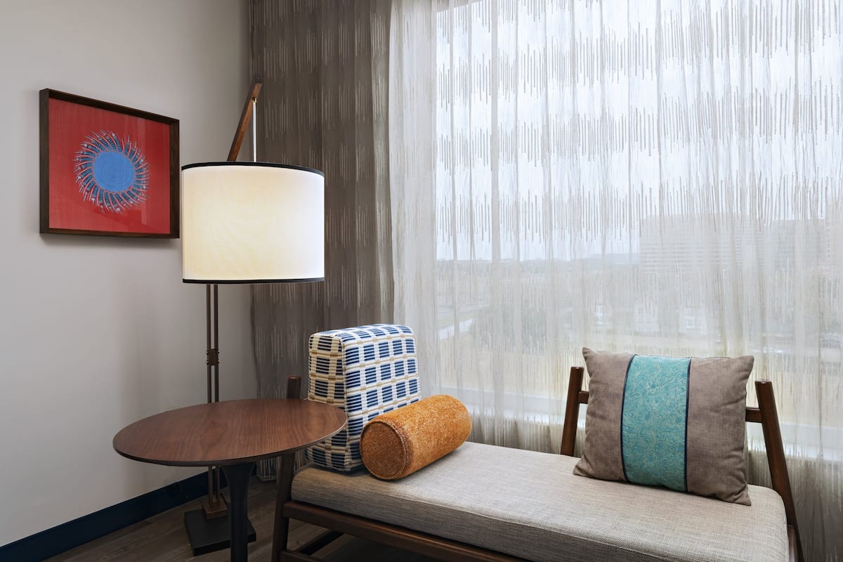 A room with a round wooden table, a floor lamp, a cushioned bench with a pillow, a framed artwork, and a window with sheer curtains.