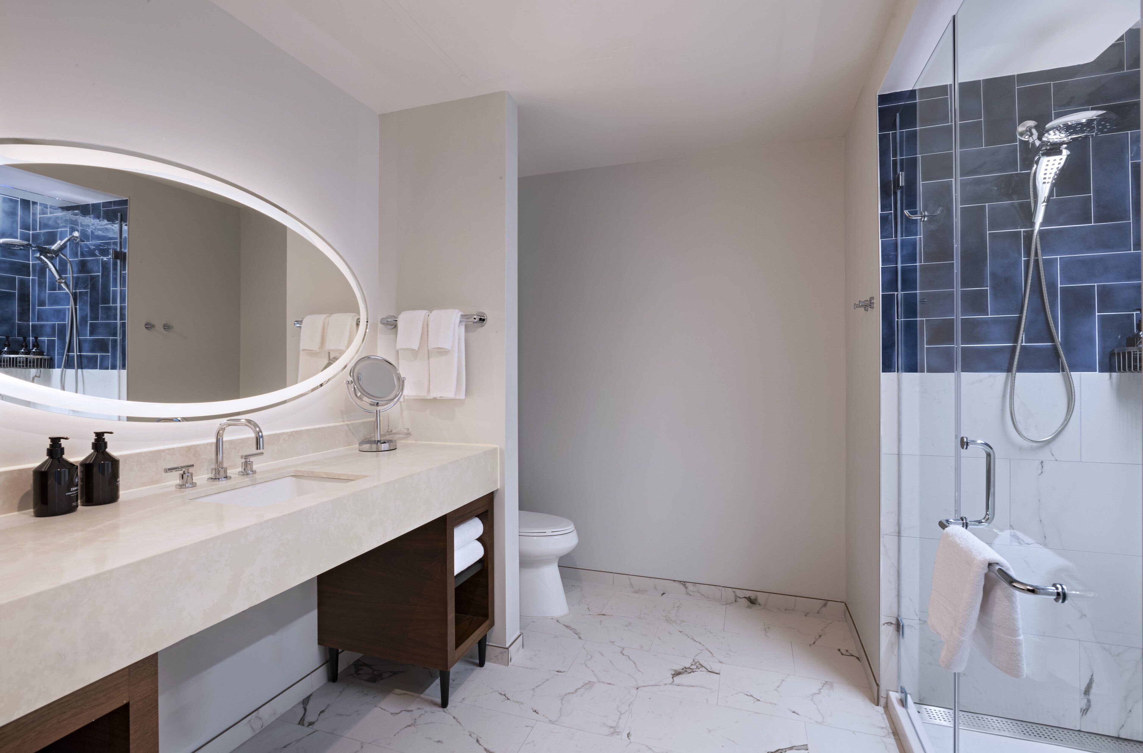 A modern bathroom with a large oval mirror, a dual sink vanity, a toilet, and a glass-enclosed shower area.