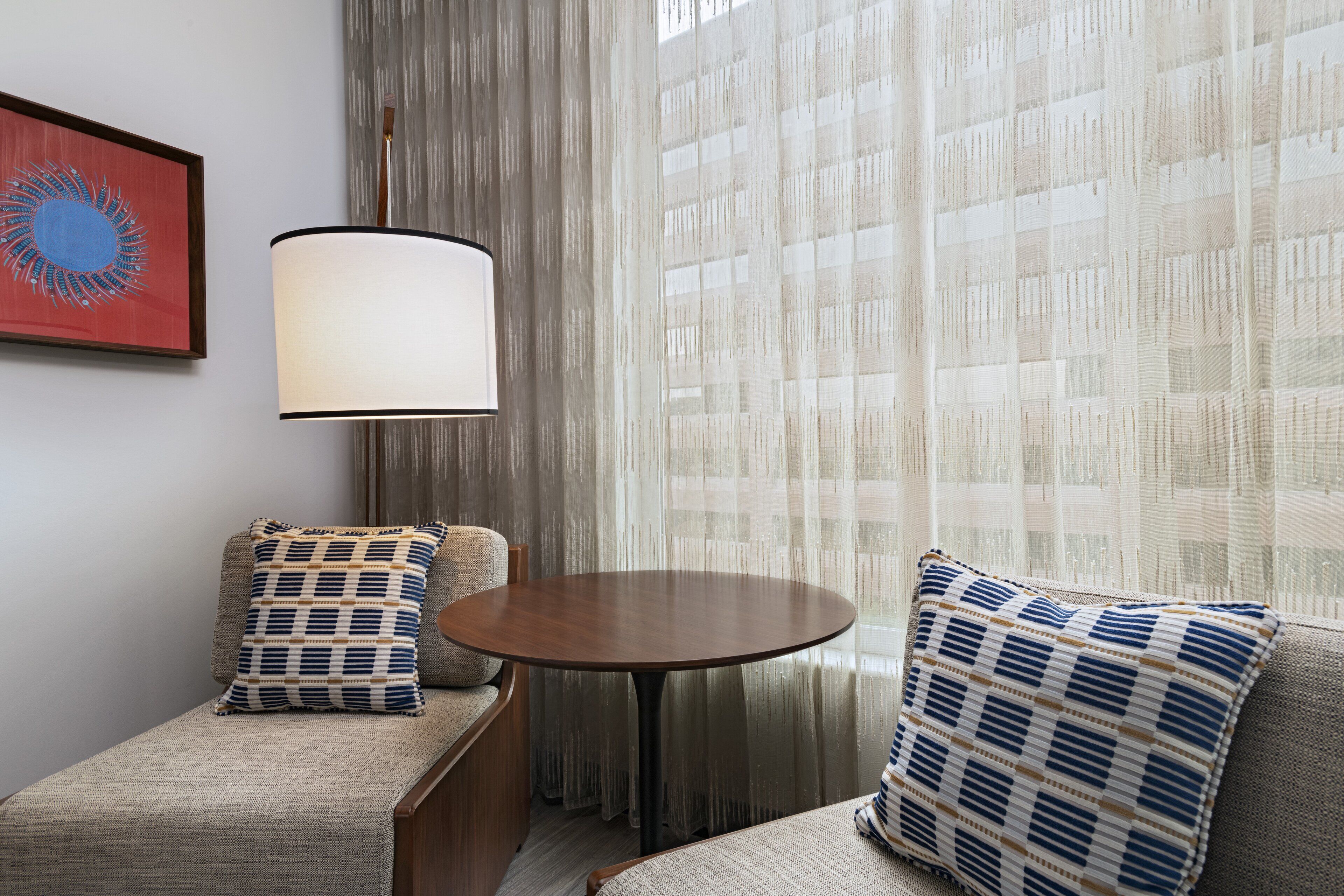 A hotel room with a round table, two sofas, a lamp, and a window with sheer curtains.