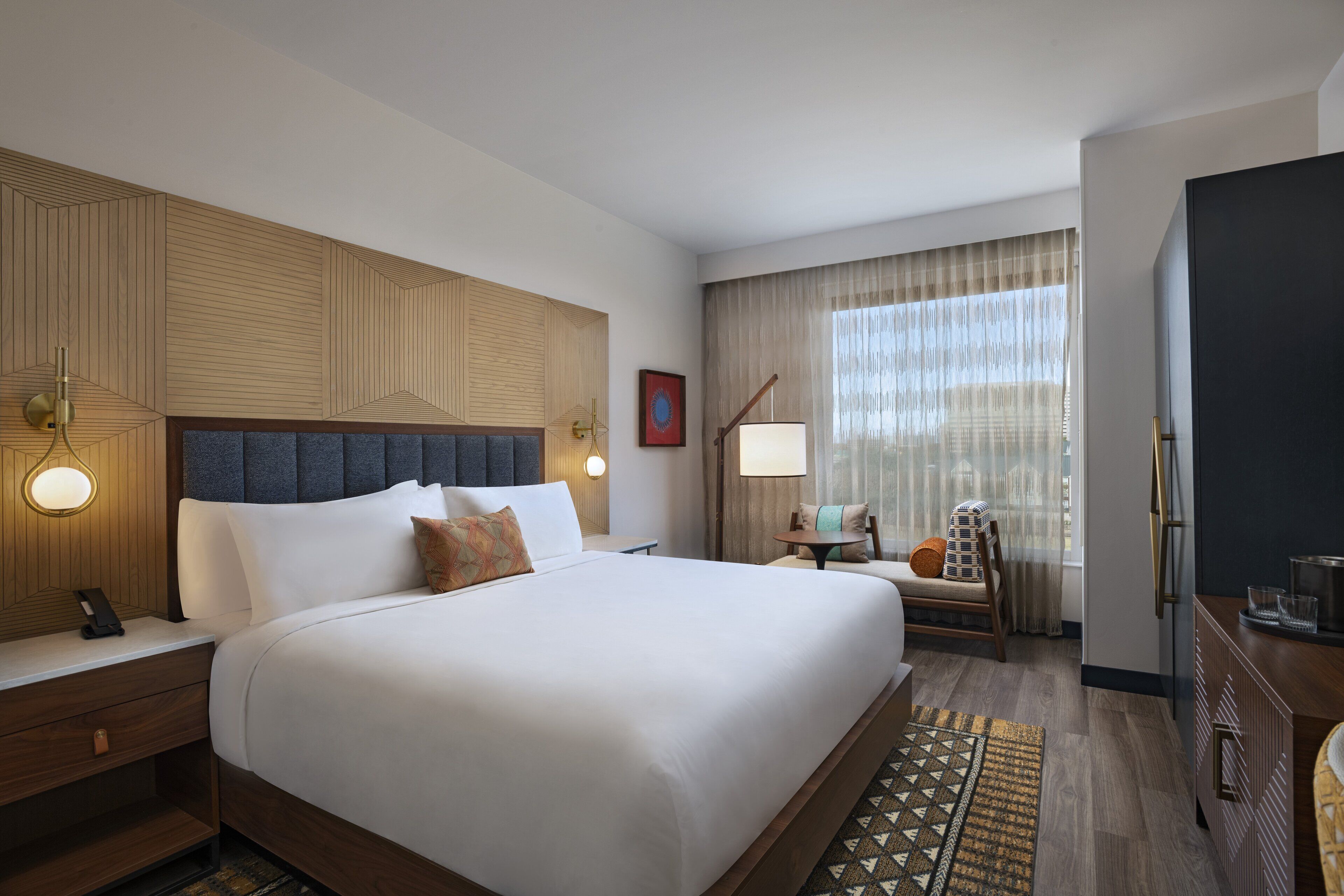 Habitación de hotel con una cama grande, cabecero de madera, mesitas de noche con lámparas, una pequeña mesa con una silla y vistas a la ciudad a través de grandes ventanales.
