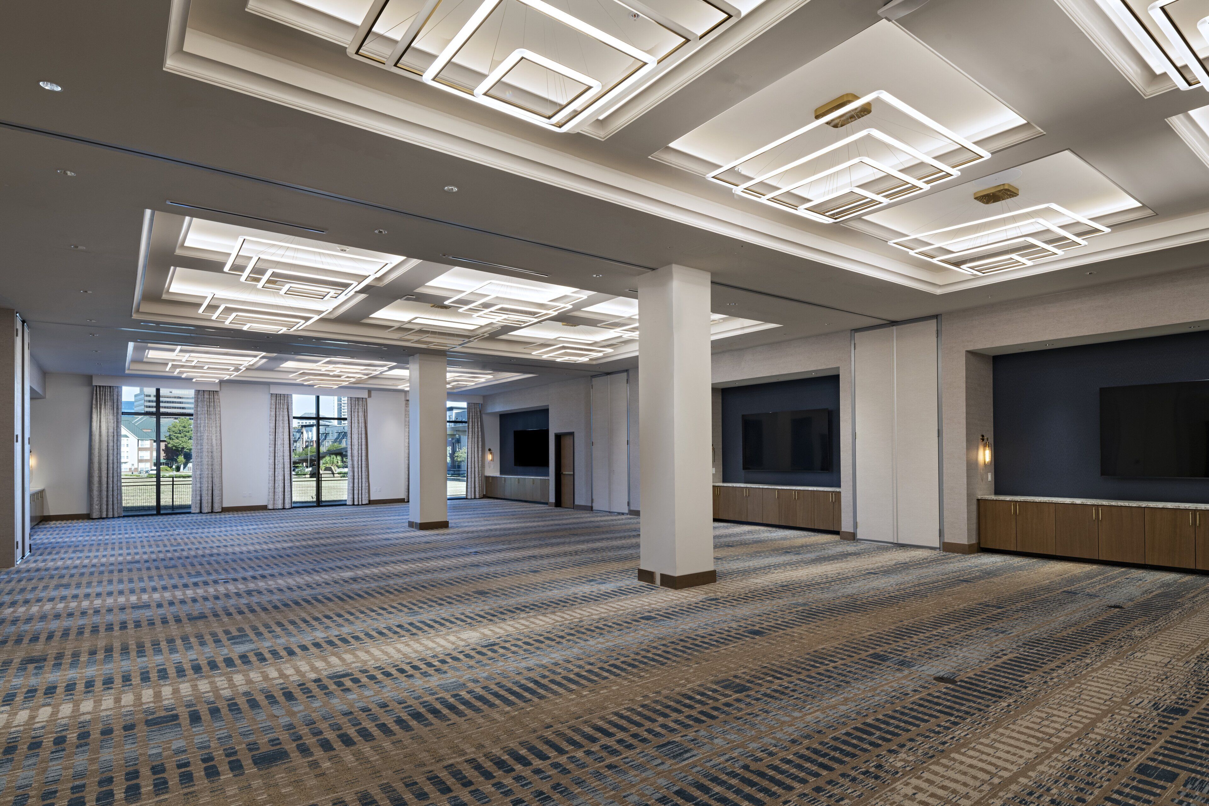 A spacious hotel lobby with a patterned carpet, large windows, and modern ceiling lighting.