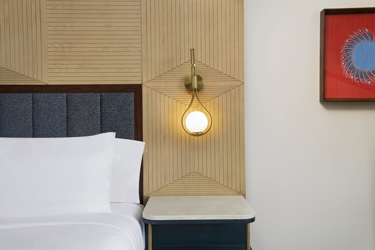 A neatly made bed with white linens, a headboard with a geometric pattern, and a wall-mounted light fixture with a round bulb.