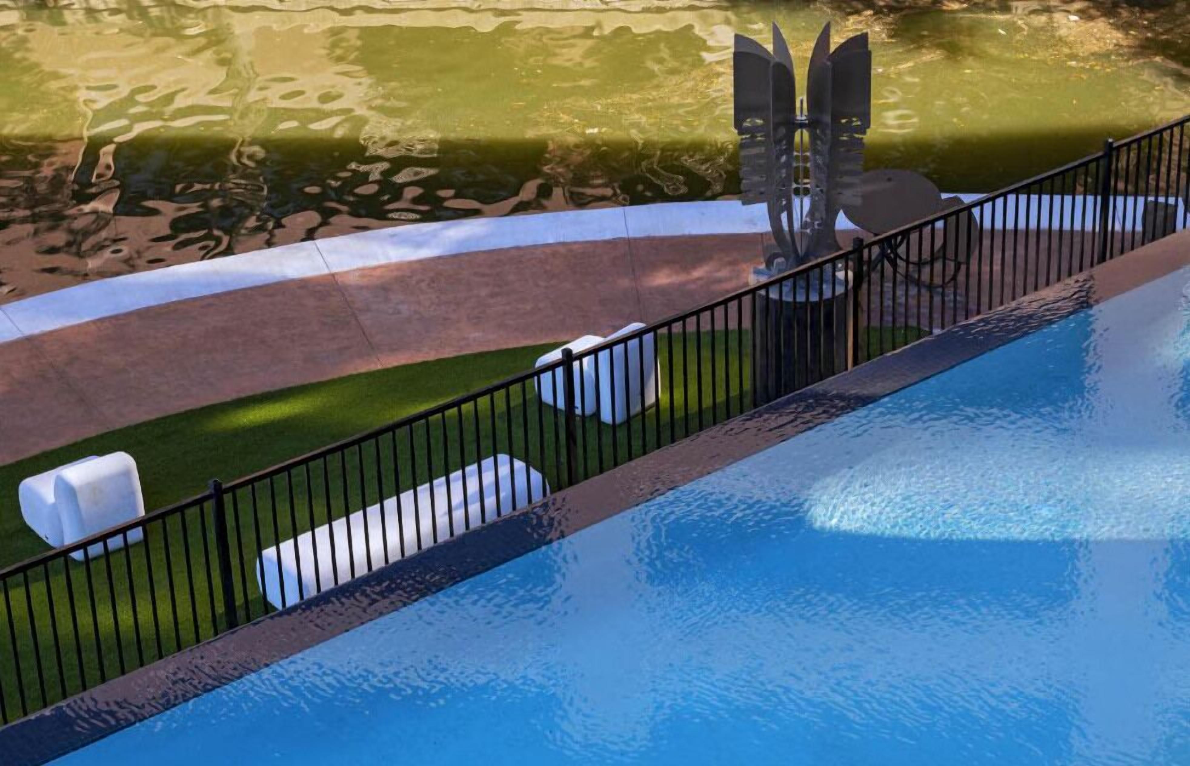 Una piscina con un borde pavimentado, una valla y un área verde con una escultura.