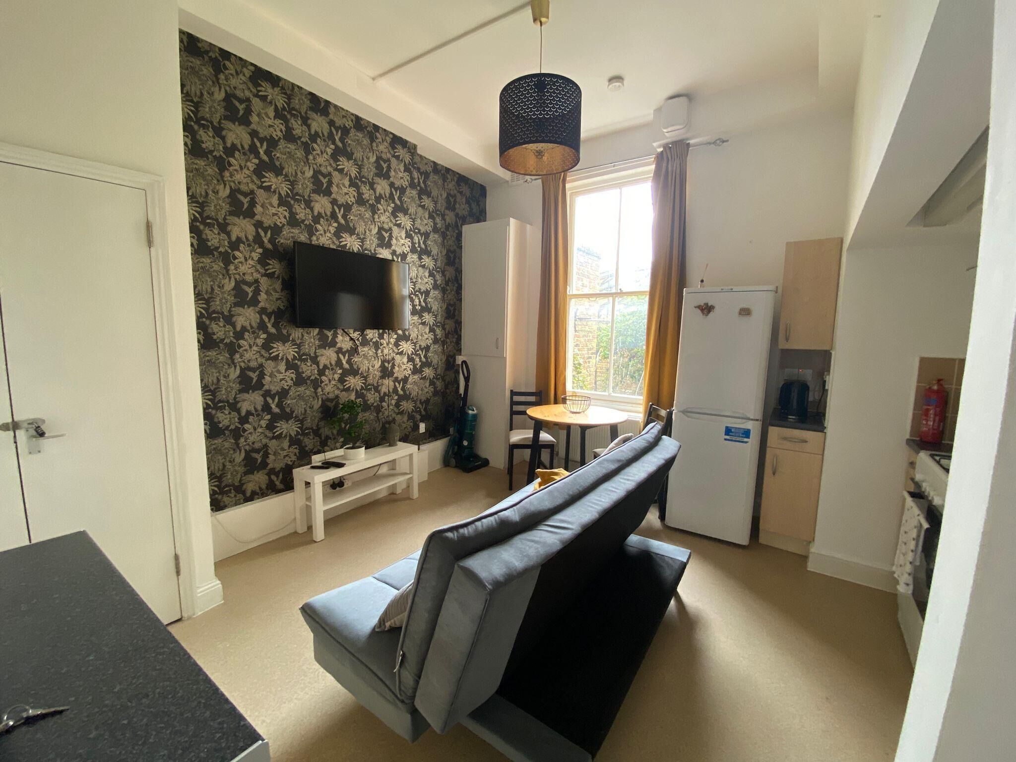 A compact living space with a sofa, a small table, a TV mounted on the wall, a refrigerator, and a window with curtains.