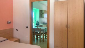 A bedroom with a bed, a wooden wardrobe, and a door leading to a kitchen area.