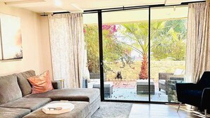 Una sala moderna con un sofá modular, un reposapiés pequeño, un ventilador de techo y un amplio ventanal corredizo que da a un área exterior con plantas y una pared amarilla.