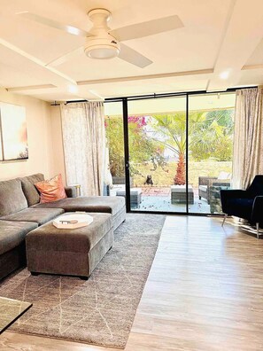A modern living room with a sectional sofa, a small ottoman, a ceiling fan, and a large sliding glass door leading to an outdoor area with plants and a yellow wall. - La Casa Maya: Modern 2bd/2br condo near Old Town. Can't beat this location!  (Scottsdale)