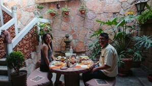 A stone-walled courtyard with a round table set for a meal, surrounded by potted plants and a decorative fountain. - HOSTAL EL REFUGIO (Puebla)