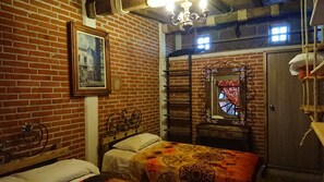 A room with a brick wall, a bed with a patterned bedspread, a wooden headboard, a hanging chair, a mirror with a decorative frame, and a window with a curtain.