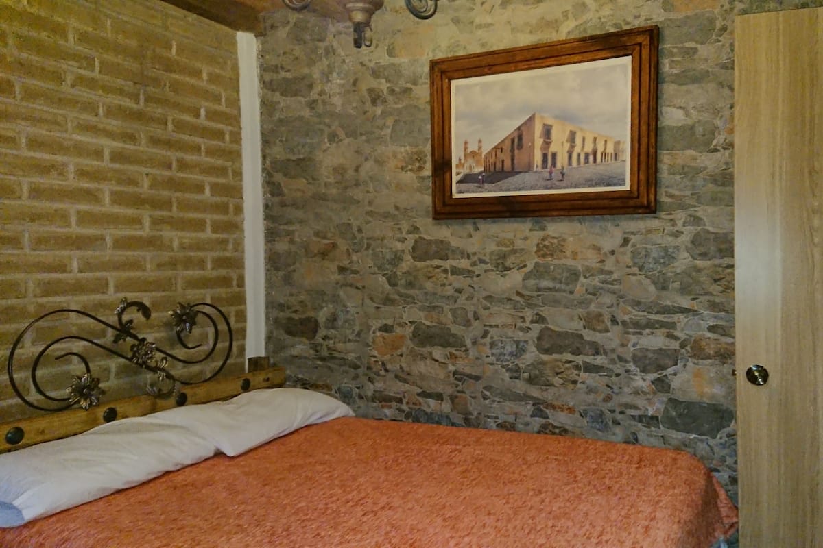 A bedroom with a stone wall, a bed with a white and orange blanket, a wooden headboard, a framed picture, and a wooden door.