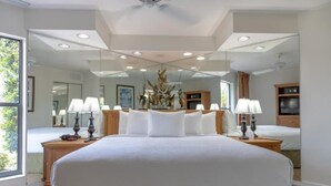 Un grand lit avec du linge de lit blanc, un miroir au-dessus et un ventilateur de plafond.