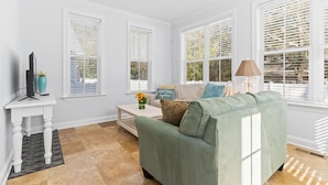A living room with a green sofa, white walls, and large windows with blinds. - 2 miles from Holden Beach Dog Friendly, Family Friendly Sandy Feet Retreat! (Supply)