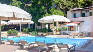 A pool area with lounge chairs and umbrellas, surrounded by greenery and a building with a red roof. - 2 bedroom cozy apartment in Vieste (Vieste)