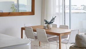 A dining area with a wooden table and white wicker chairs, a potted plant, and a view through large windows.