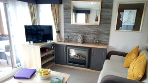 A modern living room with a flat-screen TV, a fireplace, a sofa with yellow cushions, and a dining area with a table set for two. - Lovely 2-bedroom Lodge set in Beautiful Norfolk (Great Yarmouth)
