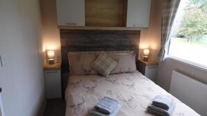 A bedroom with a bed, two bedside tables, a headboard, and a window with curtains. - Lovely 2-bedroom Lodge set in Beautiful Norfolk (Great Yarmouth)