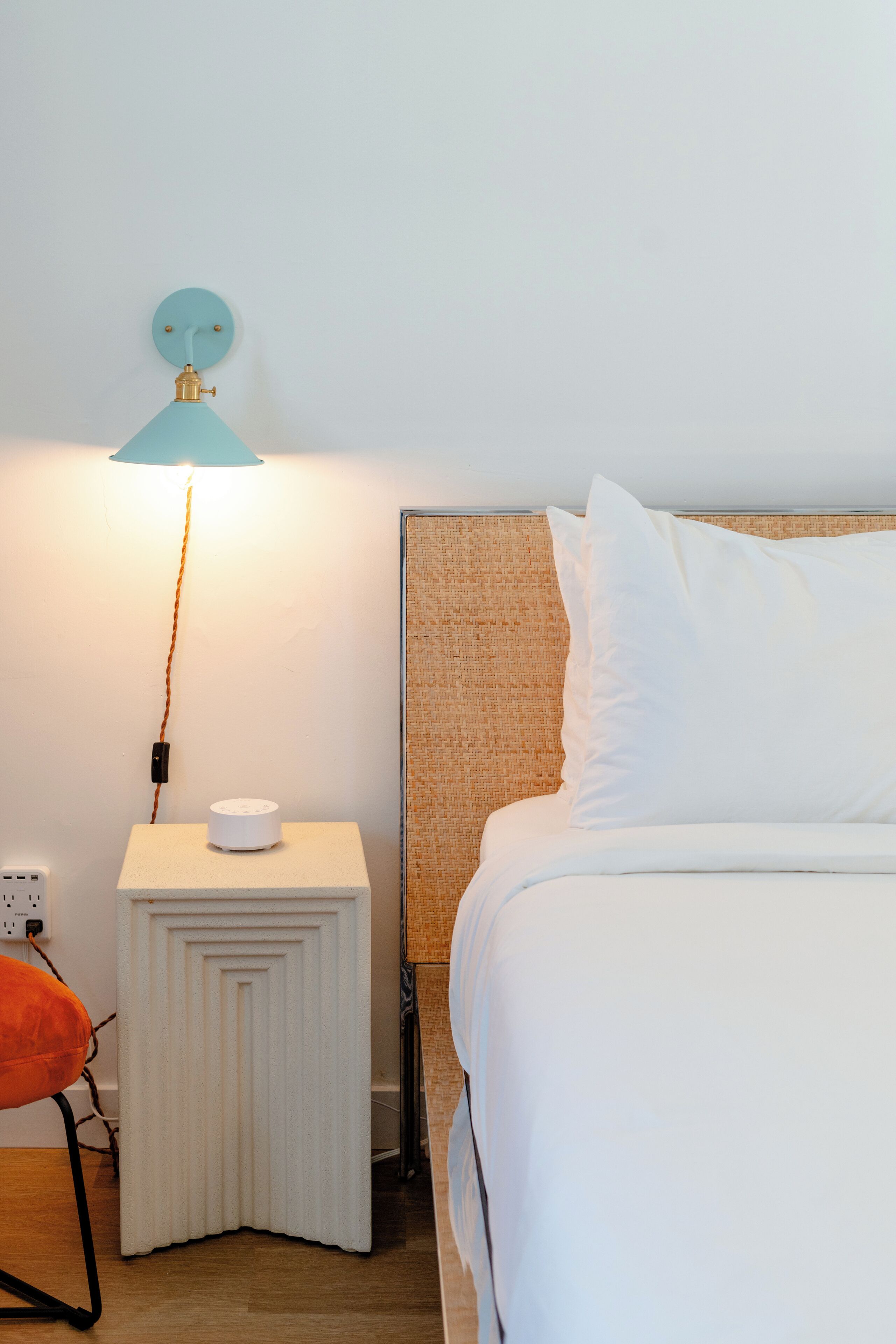 A neatly made bed with white linens, a small bedside table with a lamp, and a chair with an orange cushion.