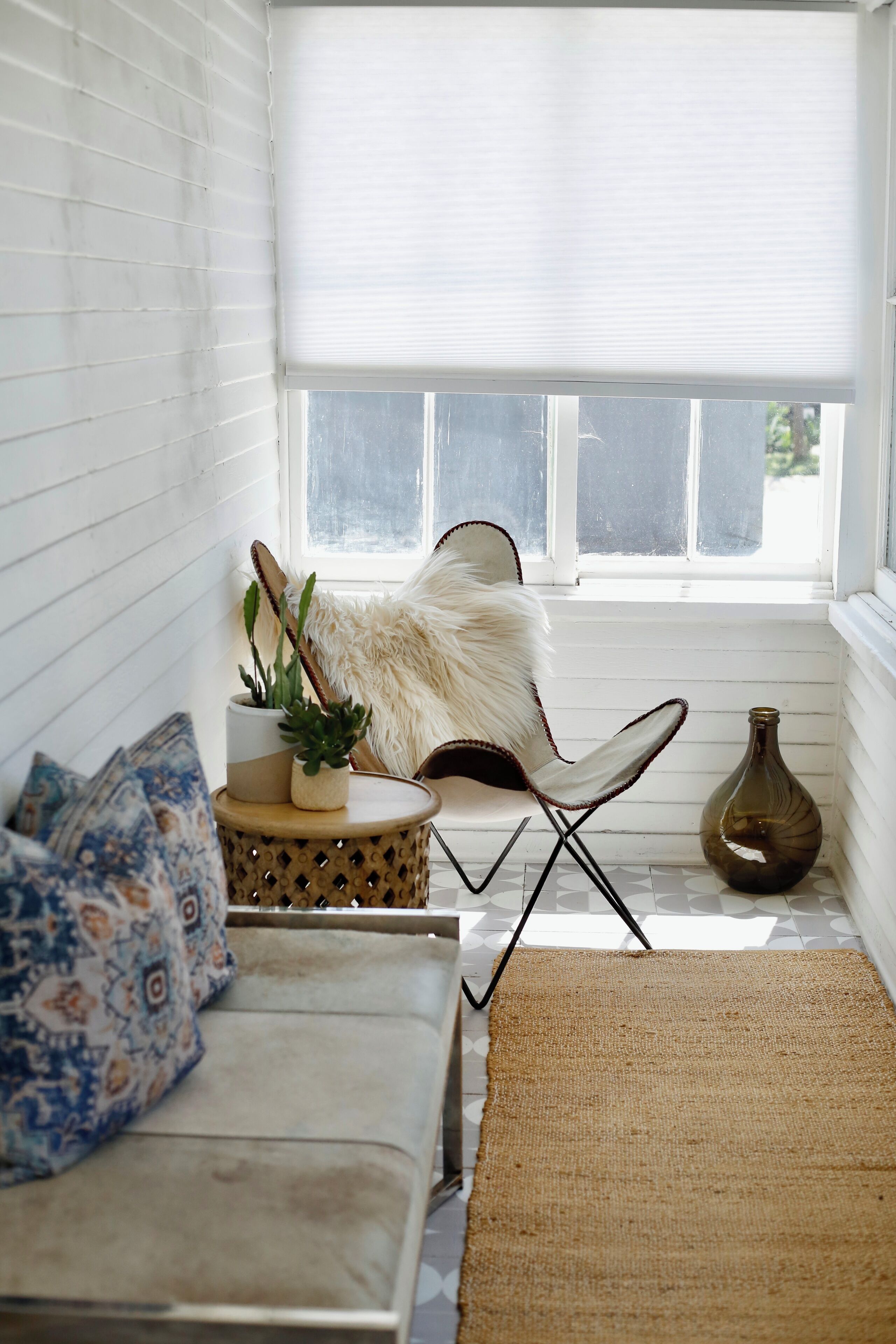 A cozy corner with a white cushioned bench, a patterned pillow, a small round table with a plant, a fluffy chair, and a vase.