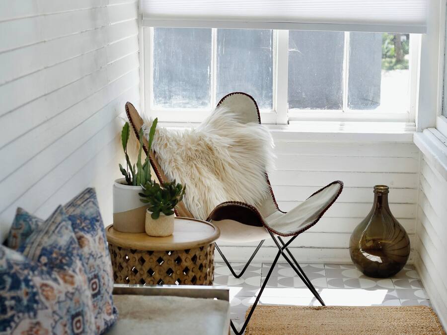 A cozy corner with a white cushioned bench, a patterned pillow, a small round table with a plant, a fluffy chair, and a vase.