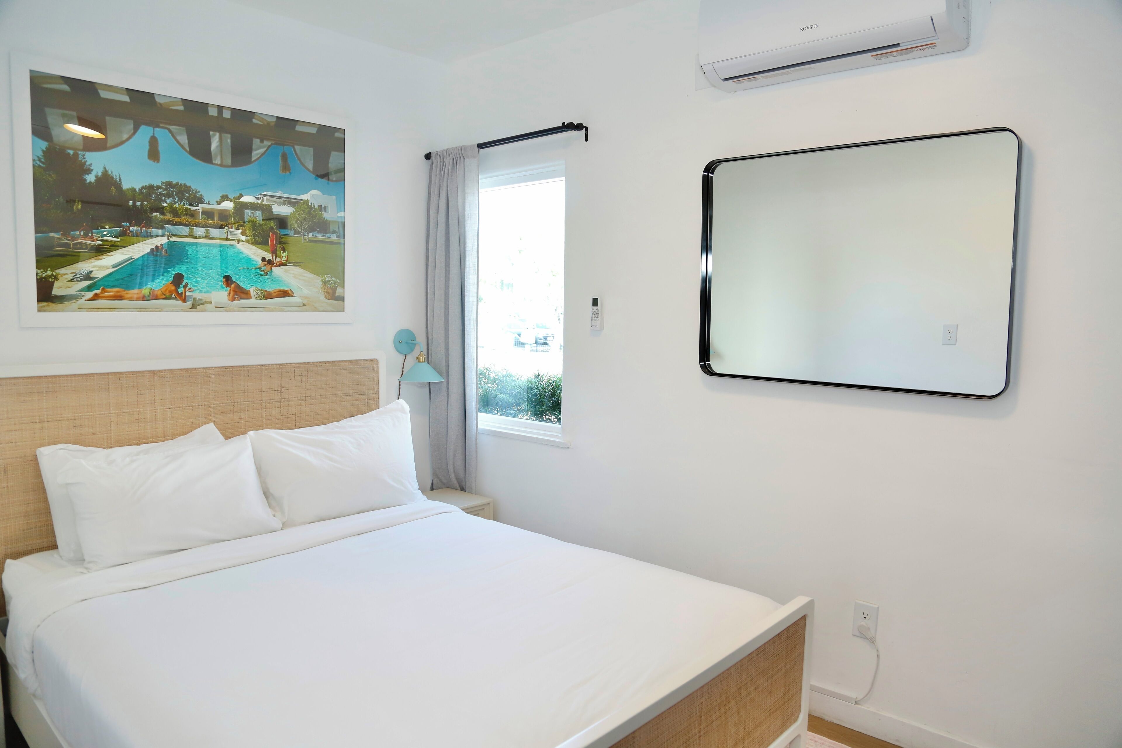 Habitación de hotel con cama, un cuadro grande enmarco con una piscina, ventana con cortina, unidad de aire acondicionado y un espejo en la pared.