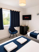 Habitación de hotel con dos camas, un televisor mostrando 'NETFLIX', una silla, una mesita con una tetera y una ventana con cortinas.