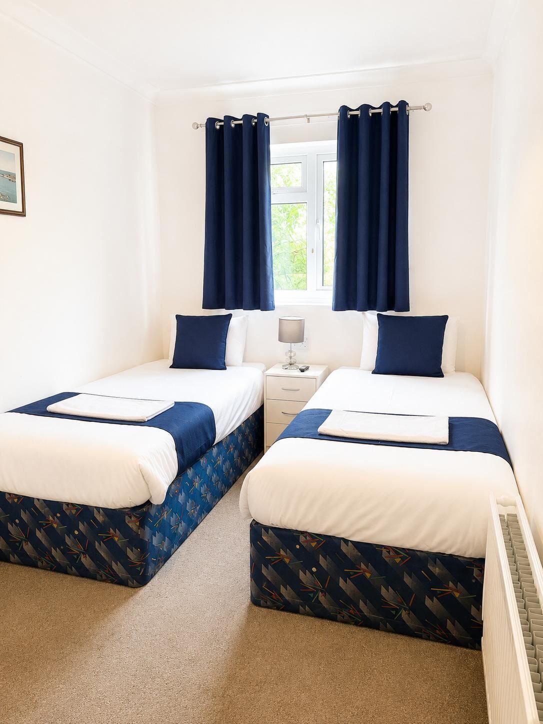 A double room with two beds, a nightstand, a lamp, and a window with curtains.