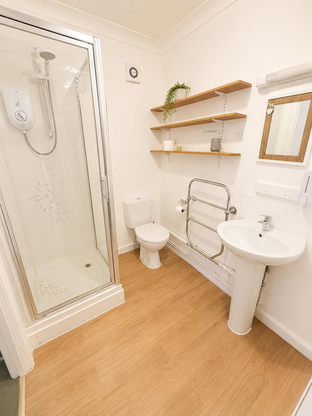 A bathroom with a glass-enclosed shower, a toilet, a sink, and wooden shelves with a plant.
