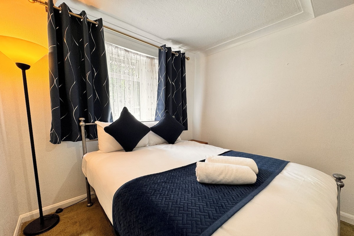 A neatly made bed with a dark blue quilt, two black pillows, and a white blanket folded at the foot. A floor lamp is positioned to the left of the bed, and a window with dark curtains is visible in the background.