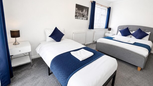A hotel room with two beds, a nightstand, a lamp, and a window with curtains. - High Trees Gatwick Guest House  (Horley)