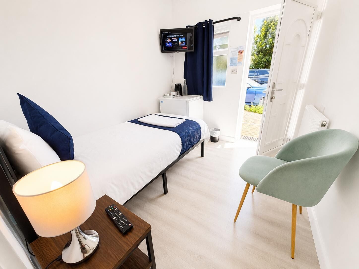 Camera d'albergo con un letto, una scrivania con una lampada, una sedia, un televisore montato a parete e una porta che dà all'esterno.