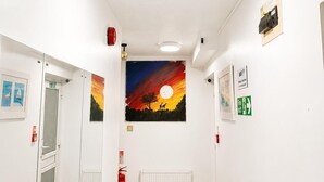 A hallway with white walls, a ceiling light, a fire extinguisher, and a painting of a sunset. - High Trees Gatwick Guest House  (Horley)