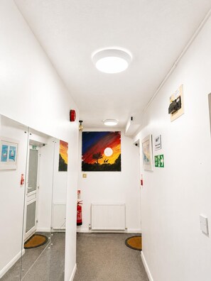 A hallway with white walls, a ceiling light, a fire extinguisher, and a painting of a sunset. - High Trees Gatwick Guest House  (Horley)