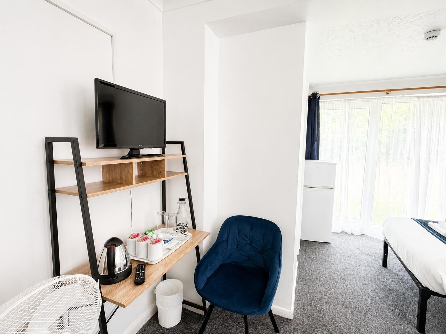 Et hotelværelse med et fjernsyn på en træhylle, en blå stol, en seng og et vindue med gardiner.