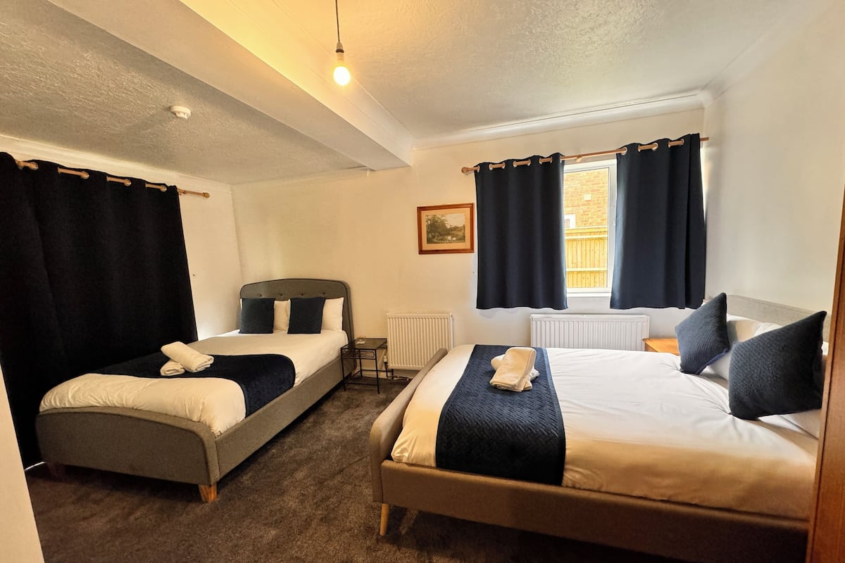 A hotel room with two single beds, a small table, a radiator, and a window with curtains.