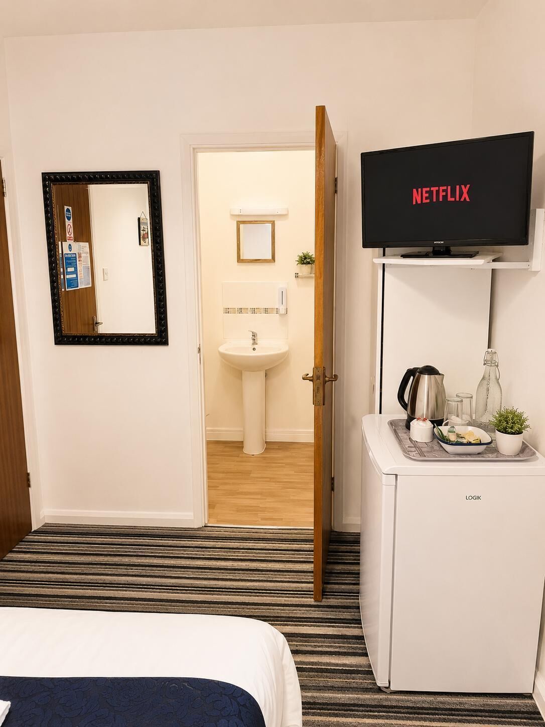 Et hotelværelse med en seng, et skrivebord med kaffemaskine, et fjernsyn der viser 'NETFLIX' og et badeværelse, der kan ses igennem en åben dør.