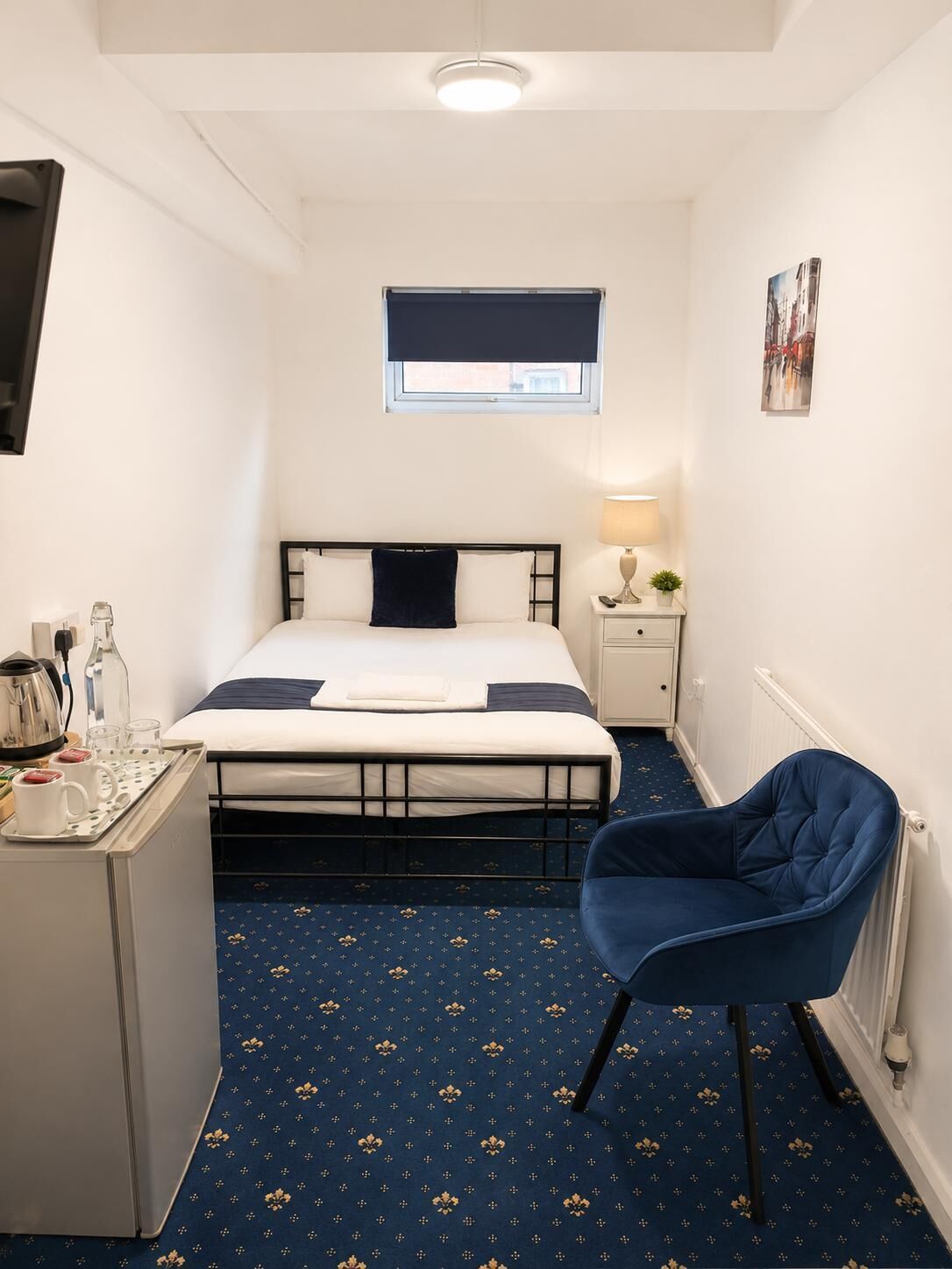A small, compact hotel room with a single bed, a desk with a kettle and cups, a chair, a small table with a lamp, and a radiator.