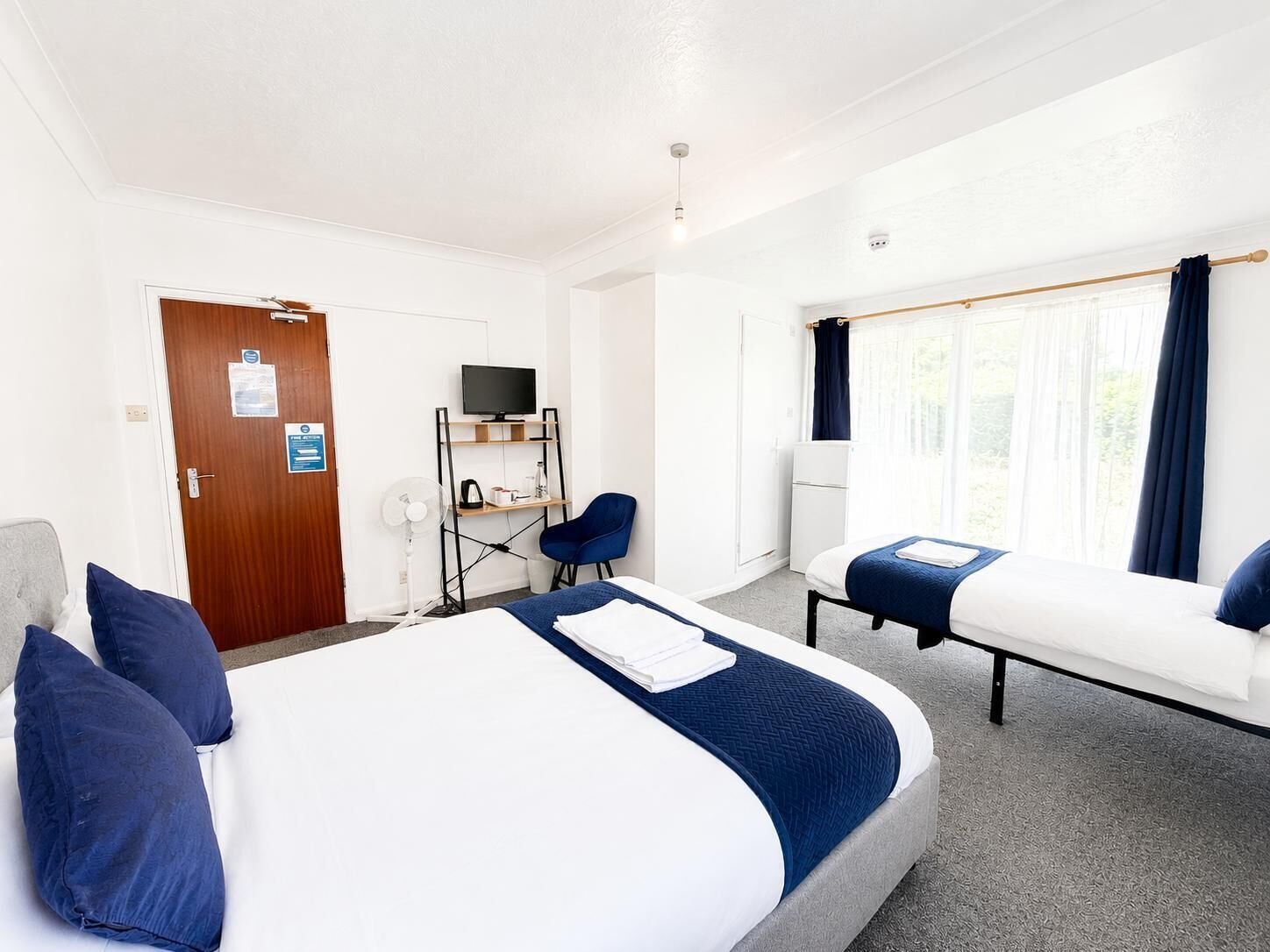 Une chambre d’hôtel avec un grand lit, un plus petit lit, un bureau avec une télévision, une chaise et une fenêtre avec des rideaux.