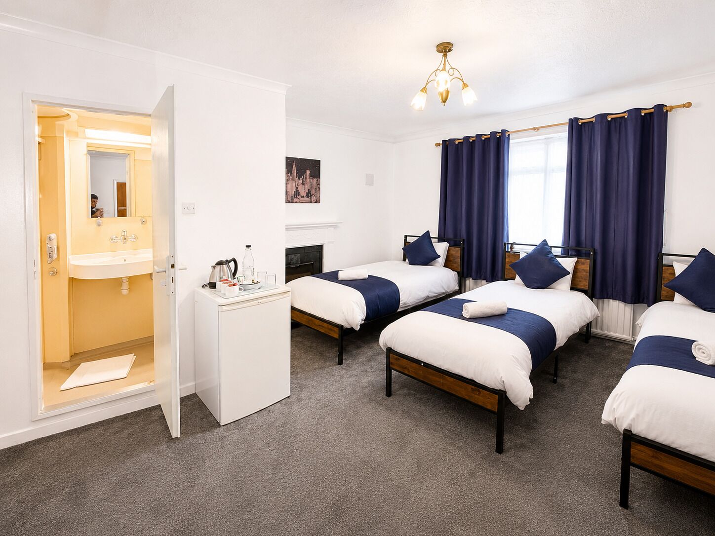 Quarto de hotel com duas camas de solteiro, um guarda-roupa branco, uma mesinha com chaleira e um quadro na parede.
