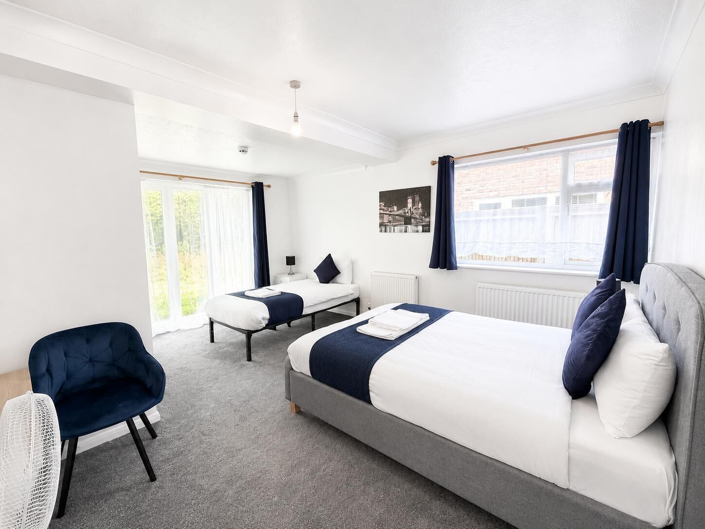 A modern hotel room with a large bed, a smaller bed, a chair, a bedside table, and a view of the city.