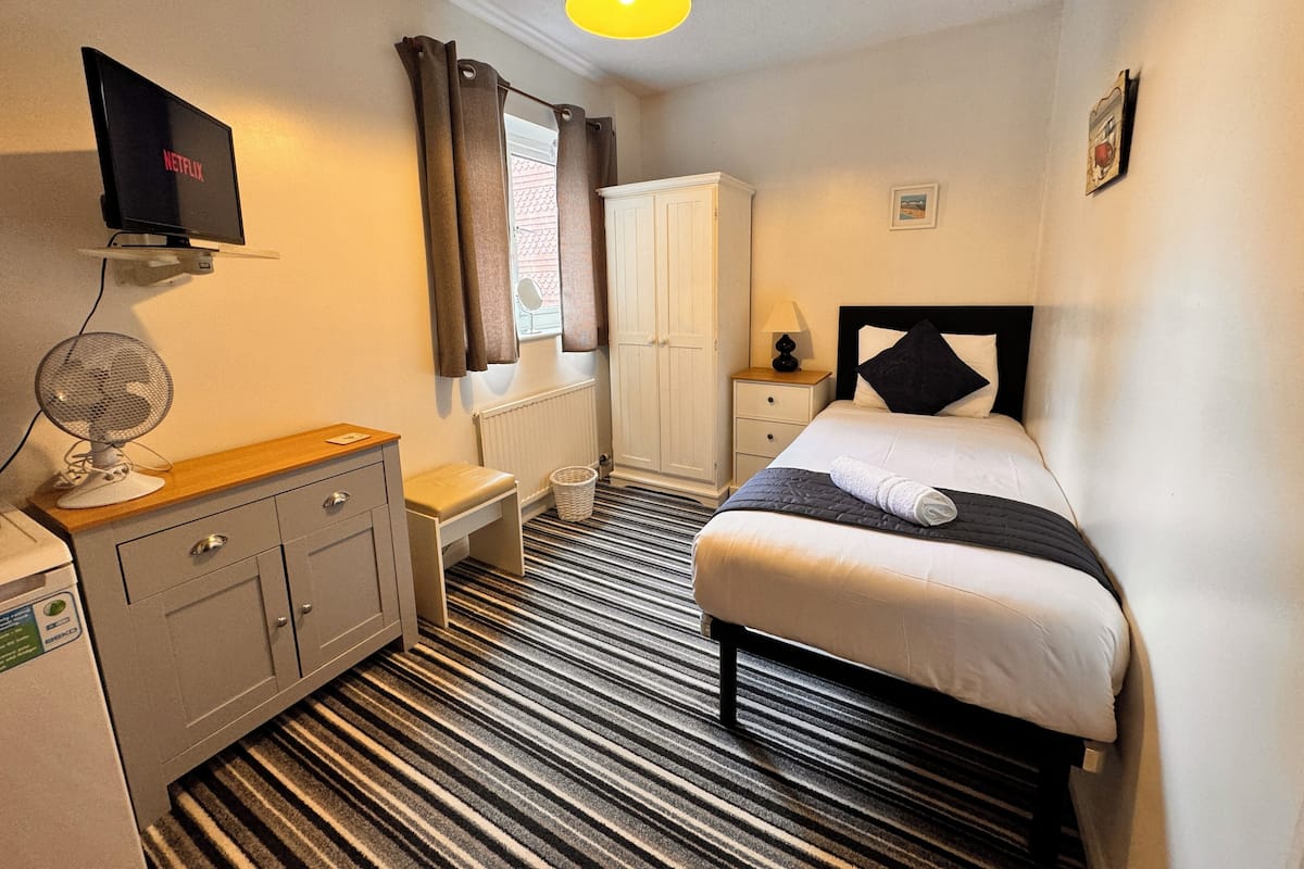 A hotel room with a bed, a desk with a computer, a wardrobe, a radiator, a window with curtains, and a ceiling light.