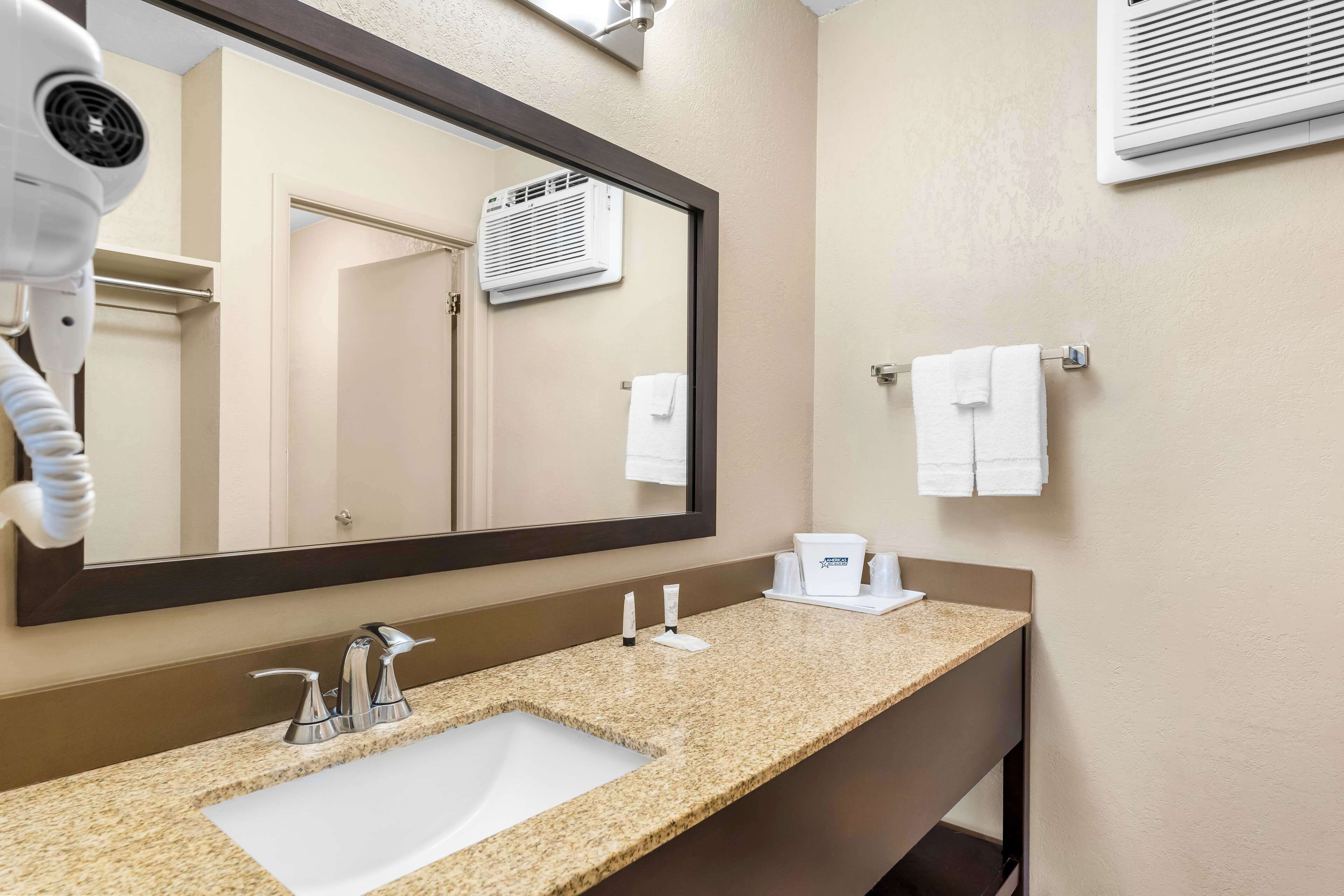 Une salle de bain d’hôtel avec un lavabo doté d’un plan de travail en granit, un grand miroir, un sèche-cheveux, un porte-serviettes et un appareil de climatisation.