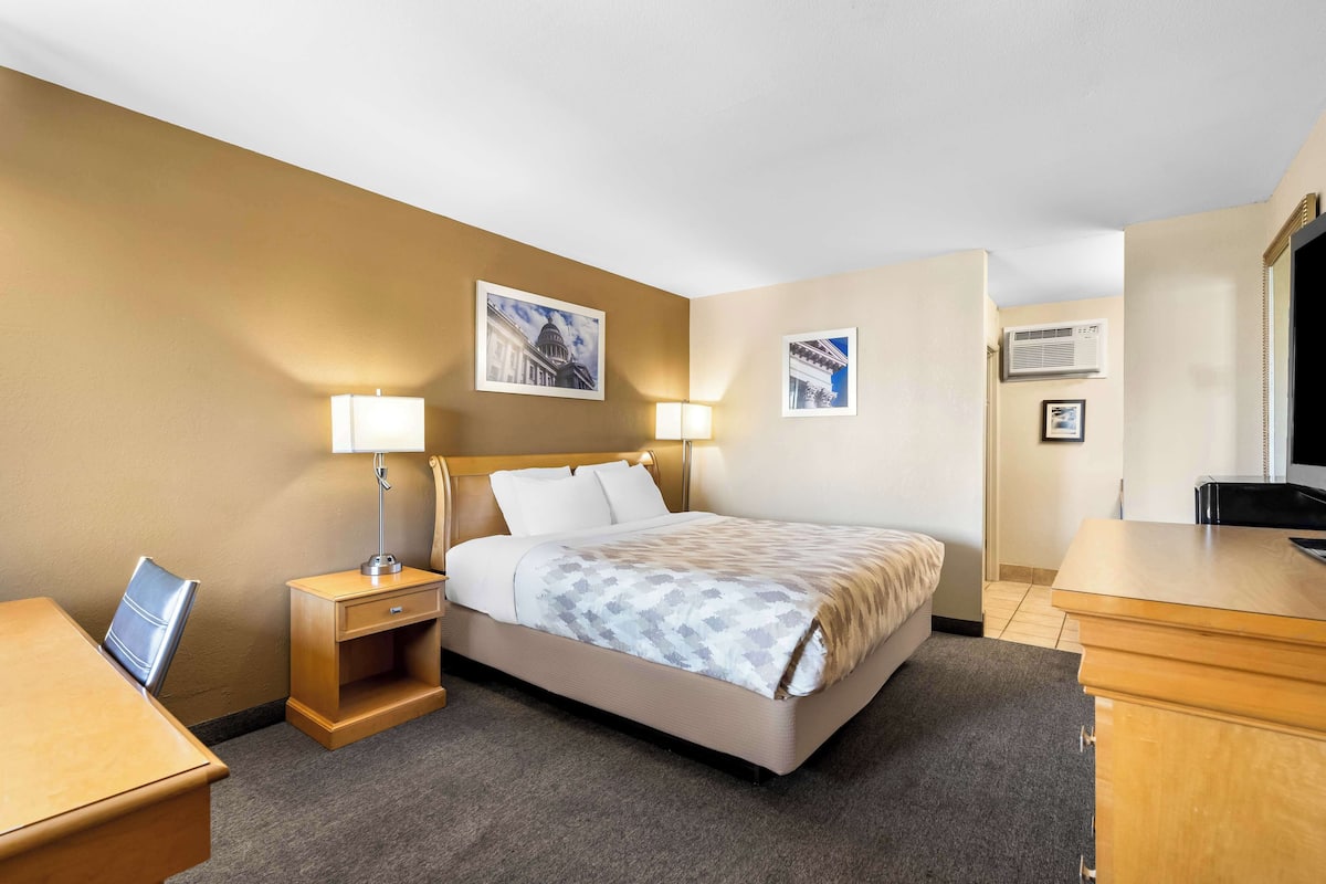 A hotel room with a bed, bedside tables, a desk, and a chair. There are wall-mounted lamps and framed pictures on the walls.