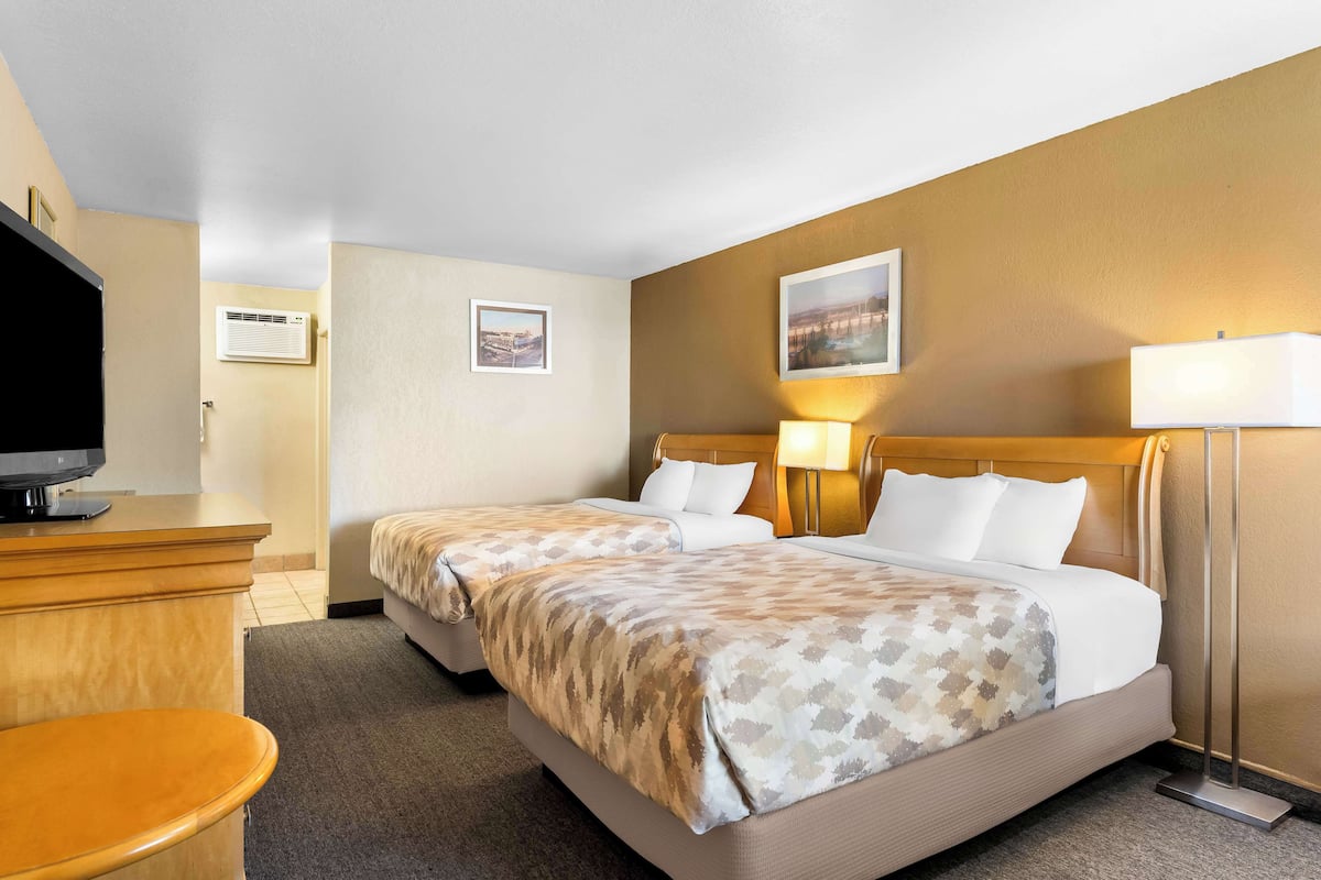 A hotel room with two beds, a desk, a television, and a wall-mounted air conditioner.