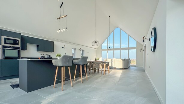 Une cuisine moderne dotée d’un îlot central, de tabourets de bar, d’un coin repas avec une table et des chaises, et de grandes fenêtres qui laissent entrer la lumière naturelle.