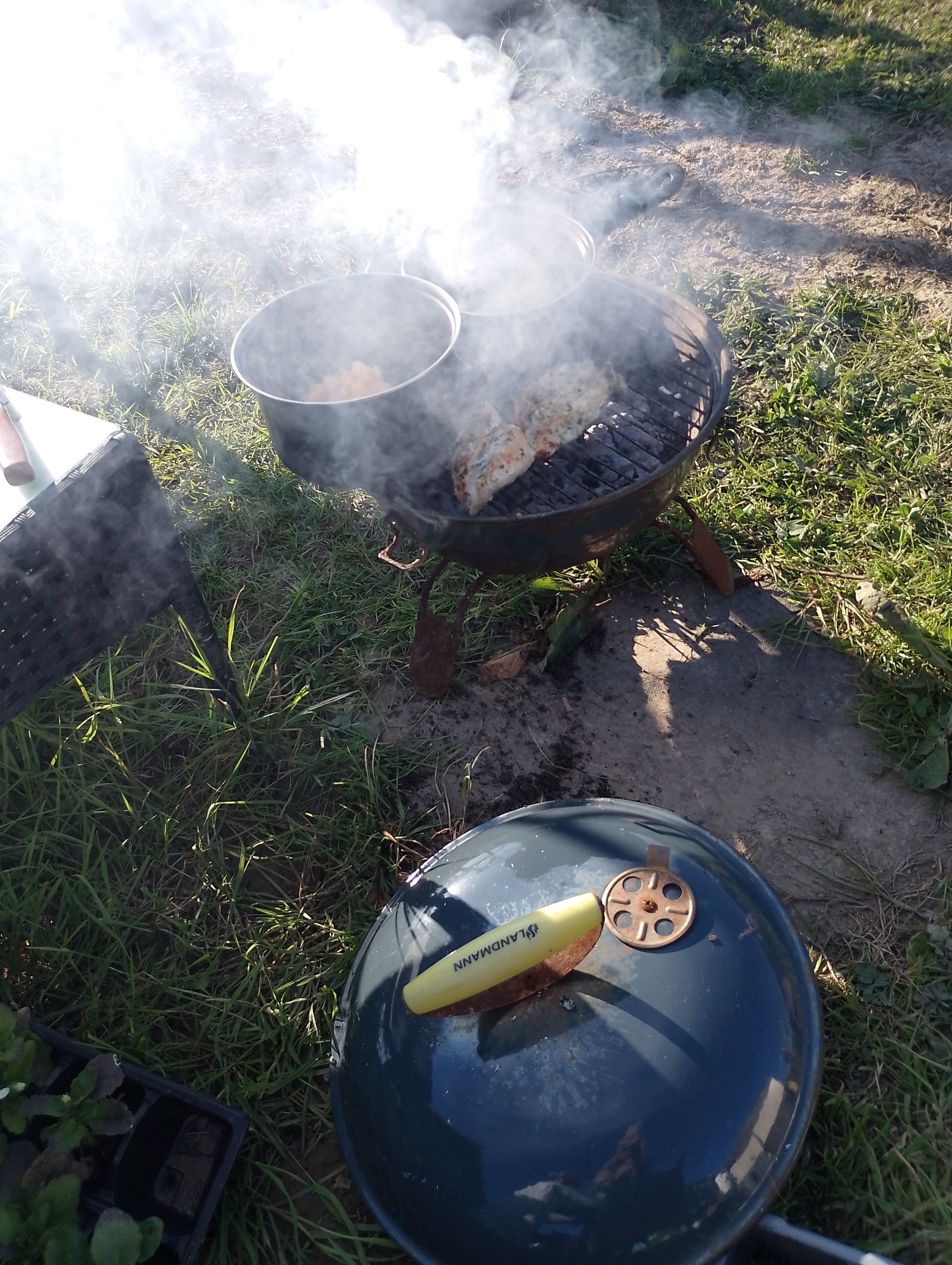 A portable grill with food cooking, a pot on a stand, and a covered grill pan on the ground.