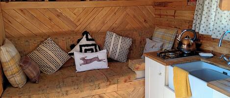 A cozy cabin interior with a herringbone-patterned floor, a wooden bench with cushions, a small kitchenette, and a rustic cabin-themed pillow.