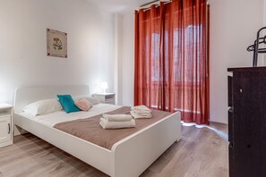 A neatly made bed with pillows and a folded towel, a bedside table with a lamp, and a red curtain on the window. - Pavone - Sweet Dreams in Florence (Firenze)