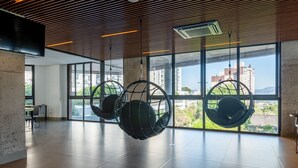 A spacious indoor area with large windows, hanging chairs, and a view of buildings and greenery outside. - LIV by LEMMON HOMES (Porto Alegre)