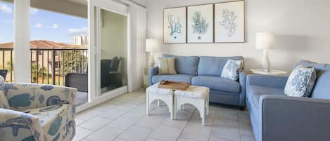 A living room with a blue sofa, a patterned armchair, and a white coffee table. There are three framed artworks on the wall and two lamps on side tables.